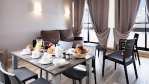 Une table de petit-déjeuner, des chaises et un canapé sont disposés dans une pièce moderne dotée de grandes fenêtres et de rideaux.