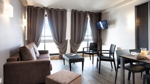 A hotel room with a sofa, two armchairs, a small table with a laptop, a TV mounted on the wall, and a breakfast setup with a glass of orange juice and a croissant. - Sleeps 6 | 1-Bedroom | Hidden Gem in Les Menuires (Les Menuires)
