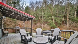 Eine Holzterrasse mit Adirondack-Stühlen und einem Tisch, umgeben von einem Wald.