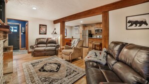 Ein großzügiges Wohnzimmer mit Ledersofas, einem gemusterten Teppich, Holzbalken und einer Küche im Hintergrund.