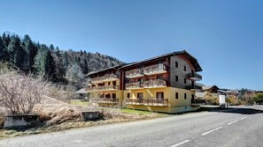 A multi-story hotel with balconies, located near a road and surrounded by forest.