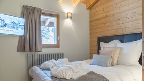 A hotel room with a bed, a window with curtains, a wall-mounted TV, and a wooden ceiling. - Praz-sur-Arly | Sleeps 6 | Never-Ending Thrills (Praz-sur-Arly)