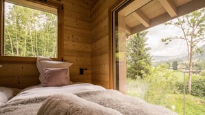 A wooden cabin bedroom with a bed, pillows, and a view of a forest and mountains. - Praz-sur-Arly | Sleeps 8 | Boundless Excitement (Praz-sur-Arly)