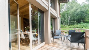A wooden deck with outdoor furniture, a glass door leading to an interior dining area, and a view of a forested area. - Praz-sur-Arly | Sleeps 8 | Boundless Excitement (Praz-sur-Arly)