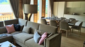A cozy living room with a sofa, dining table, and chairs. There is a kitchen area with cabinets and a window.