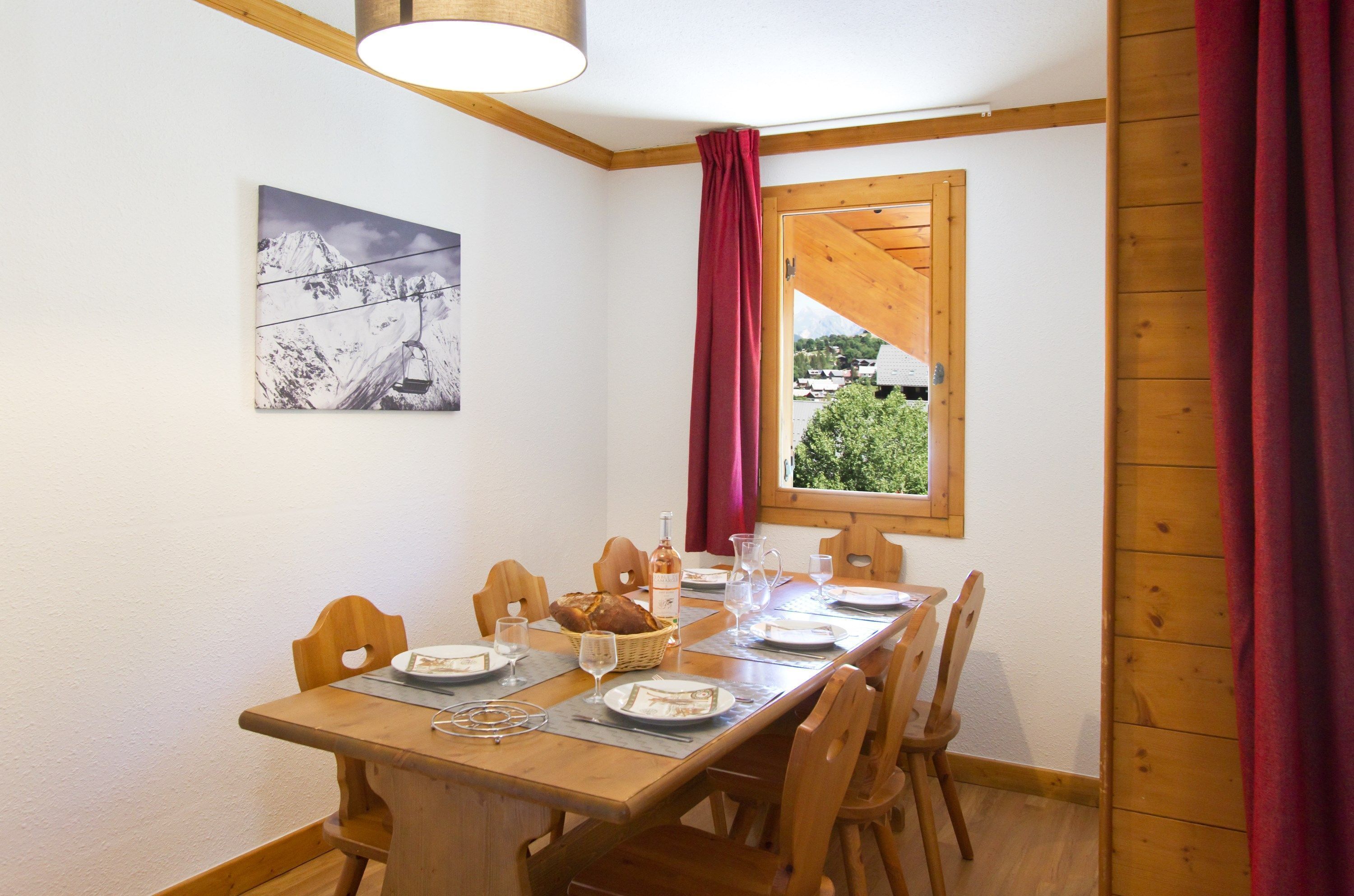 Un coin repas avec une table en bois dressée pour quatre personnes, des chaises, une fenêtre donnant sur un paysage verdoyant et un tableau accroché au mur.