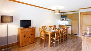 A dining area with a wooden table and chairs, a television on a cabinet, and a kitchen in the background.