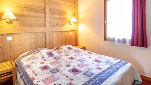 A bedroom with a wooden headboard, two bedside lamps, a quilted bedspread, and a window with a curtain. - Sleeps 7! 2BR Apt| Scenic Living in Valmorel! (La Léchère)