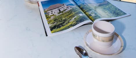 A table with a white cup and saucer, a spoon, a potted plant, and two open magazines with landscape photos.