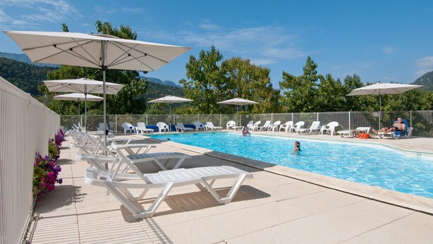 A swimming pool with white lounge chairs and umbrellas, surrounded by greenery and mountains.