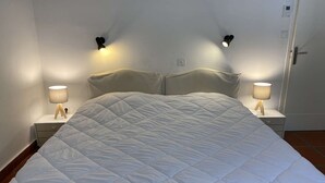 A neatly made bed with a white quilt, two bedside lamps, and a door with a handle. - Maison Familiale Proche Plage (PYLA SUR MER)