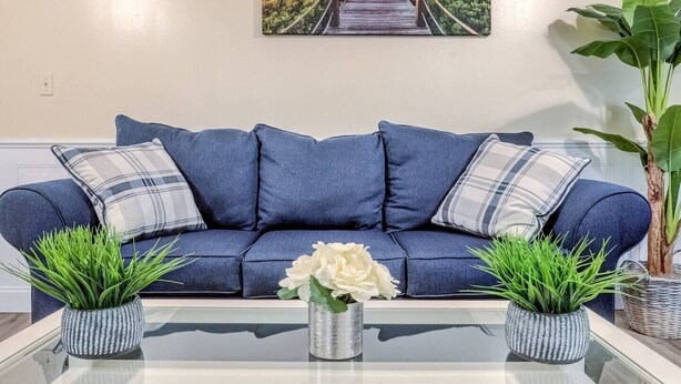 A blue sofa with plaid pillows, a glass coffee table with a white flower centerpiece, and two potted plants. - Premier League Pad - with Free Parking, Heat & AC (Manchester)
