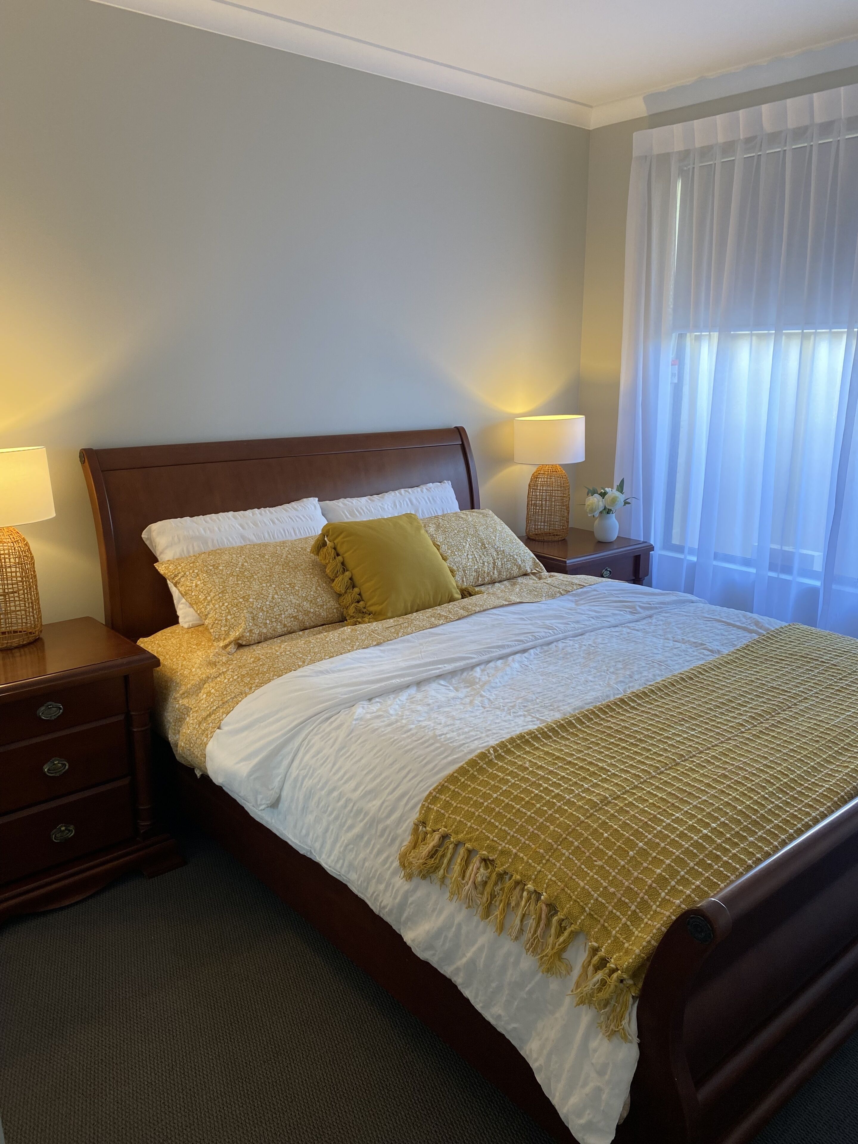 A neatly made bed with a wooden headboard, two bedside tables with lamps, a window with sheer curtains, and a small vase with flowers on the nightstand.
