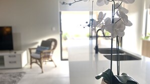 A modern living room with a white orchid plant on a metal stand, a wicker chair, a flat-screen TV, and a glass door leading outside. - New 5 Bedroom Home 1km from the Beautiful Busselton Beaches  (Geographe)