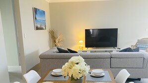A dining area with a wooden table set for four, surrounded by white chairs, with a grey sofa and a TV in the background. - New 5 Bedroom Home 1km from the Beautiful Busselton Beaches  (Geographe)