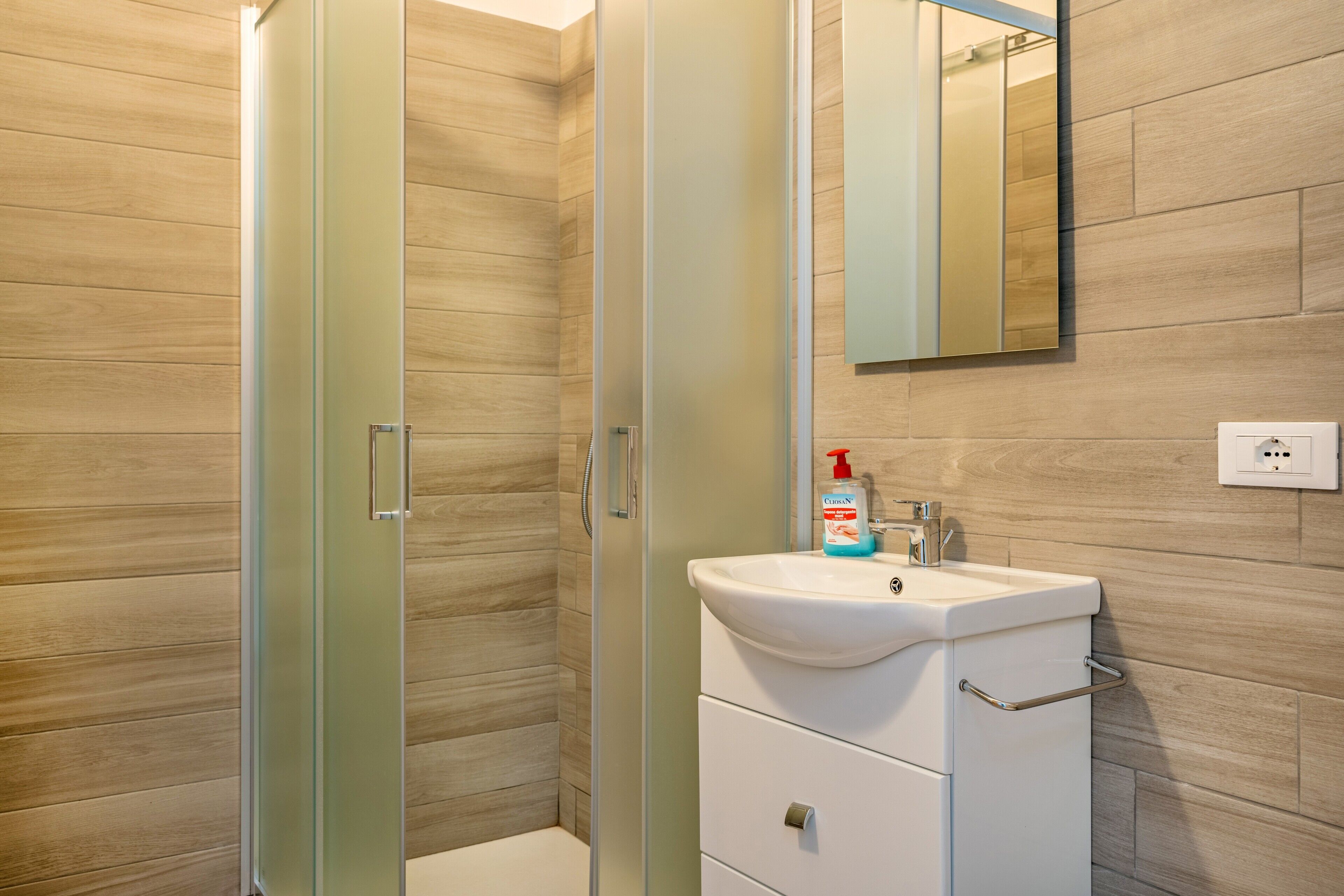 A bathroom with a glass shower enclosure, a white sink with a mirror above it, and a wooden wall.