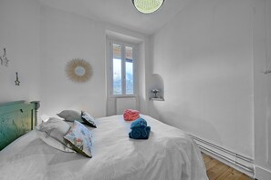 Un dormitorio con cama, ventana, espejo montado en la pared y una pequeña estantería.