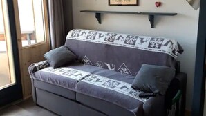 A grey sofa with patterned cushions, a framed picture above it, a small shelf, and a window with curtains.