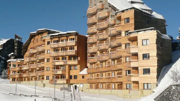 A multi-story hotel with balconies, a ski slope in the background, and snow on the roof.