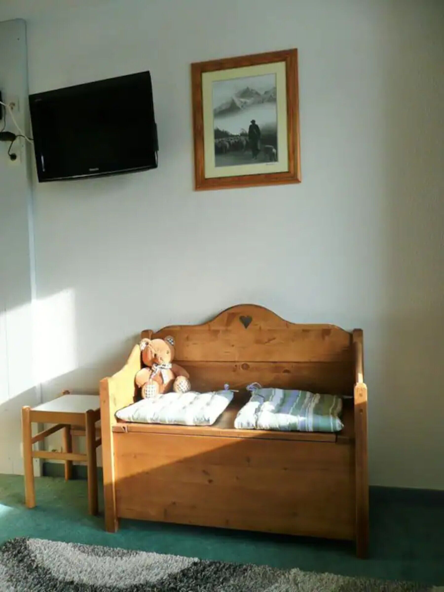 A wooden bed with pillows, a teddy bear, a framed picture, and a television on the wall.