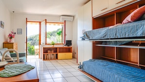 A compact room with a bunk bed, a desk with a chair, a small table, and a view of greenery through an open door.