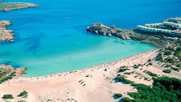 Vue aérienne d’une zone côtière comprenant une plage, des affleurements rocheux et un complexe hôtelier composé de plusieurs bâtiments.