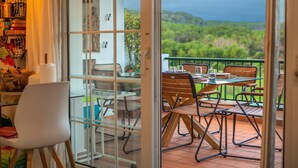 Une terrasse ouverte avec une table et des chaises, une vue sur la verdure et une porte vitrée donnant sur une pièce avec des étagères et un fauteuil.