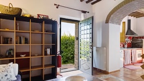 A living room with a bookshelf, a bowl on a table, a door leading to a kitchen, and a view of greenery outside. - Holiday Home 'La Ripa - Appartamento I Nidi' with Shared Pool, Garden & Wi-Fi (San Gimignano)