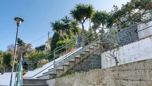 A set of outdoor stone stairs with metal railings, leading up a slope with trees and a building in the background.