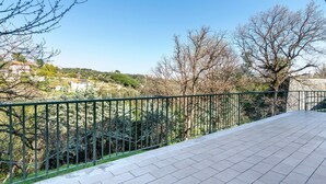 A balcony with a tiled floor, a metal railing, and a view of a hilly landscape with trees and buildings.