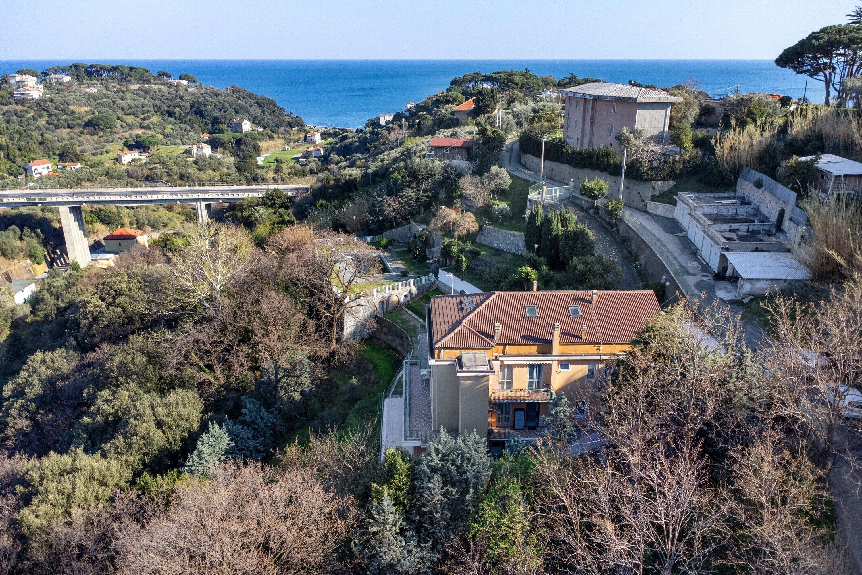 A coastal residential area with a prominent building featuring a red roof, surrounded by dense vegetation and a bridge in the background.