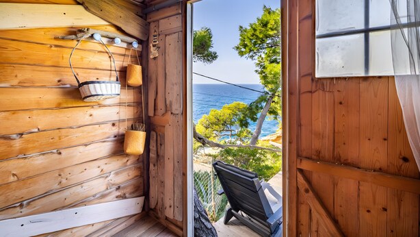 A wooden cabin interior with a view of the sea, a wooden bench, and a hanging basket.