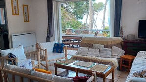 A furnished living room with a sofa, chairs, a coffee table, a TV, and a large window with a view of trees and a patio. - Holiday Home 'Villa Calanques' with Sea View, Private Terrace and Wi-Fi (Ensuès-la-Redonne)
