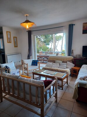 A furnished living room with a sofa, chairs, a coffee table, a TV, and a large window with a view of trees and a patio. - Holiday Home 'Villa Calanques' with Sea View, Private Terrace and Wi-Fi (Ensuès-la-Redonne)