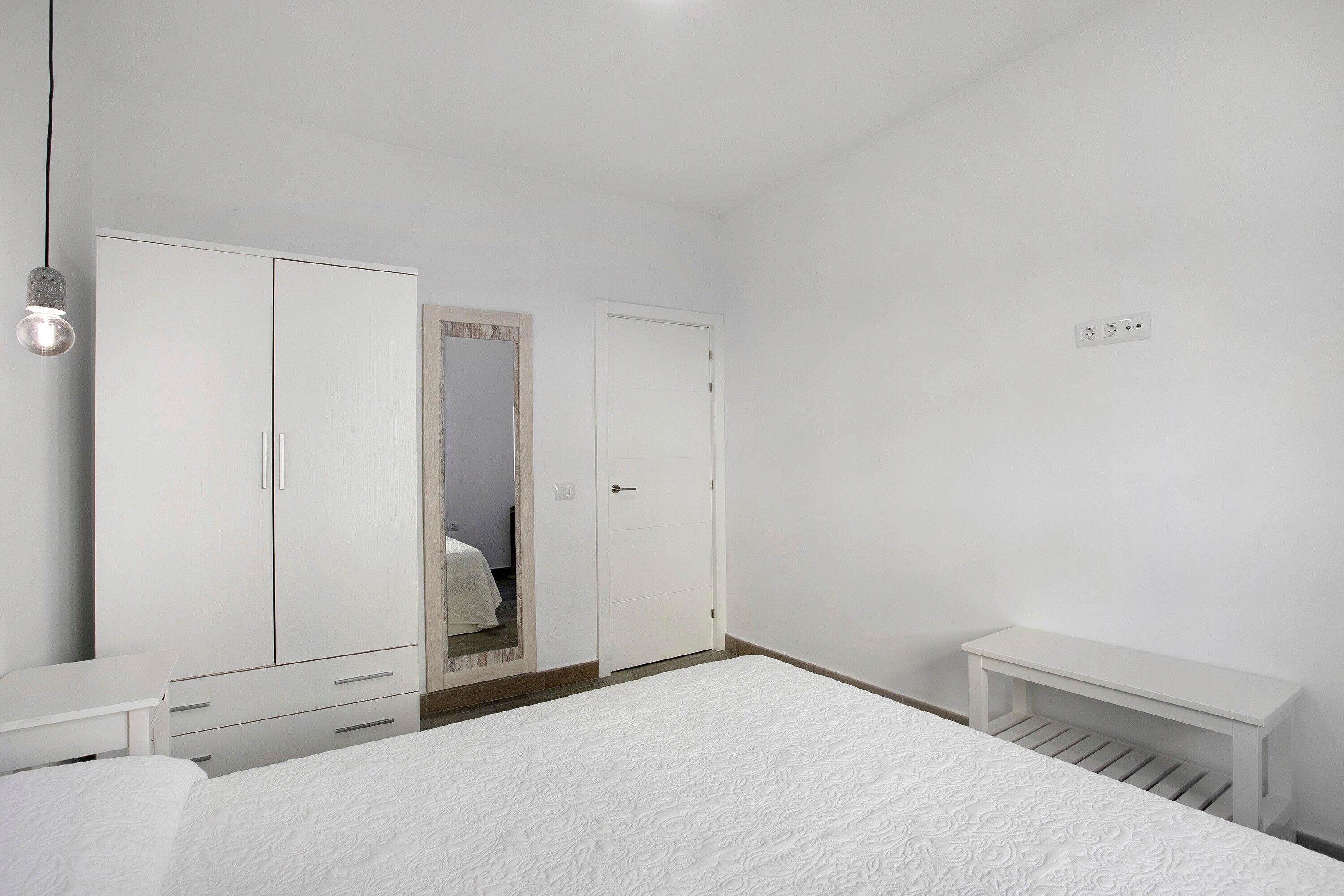 Un dormitorio con cama, armario, espejo, mesita de noche y un pequeño banco.