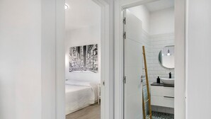 Une chambre à coucher avec un lit, un mur blanc orné d’une œuvre d’art en noir et blanc, une porte blanche et une salle de bain avec un lavabo et un miroir blancs.