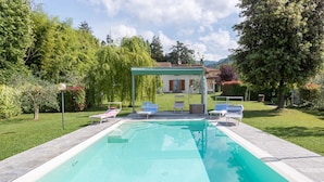 Une piscine rectangulaire à l’eau d’un bleu limpide, entourée d’une terrasse dallée. On y trouve des chaises longues et un espace salon couvert agrémenté d’une pergola.