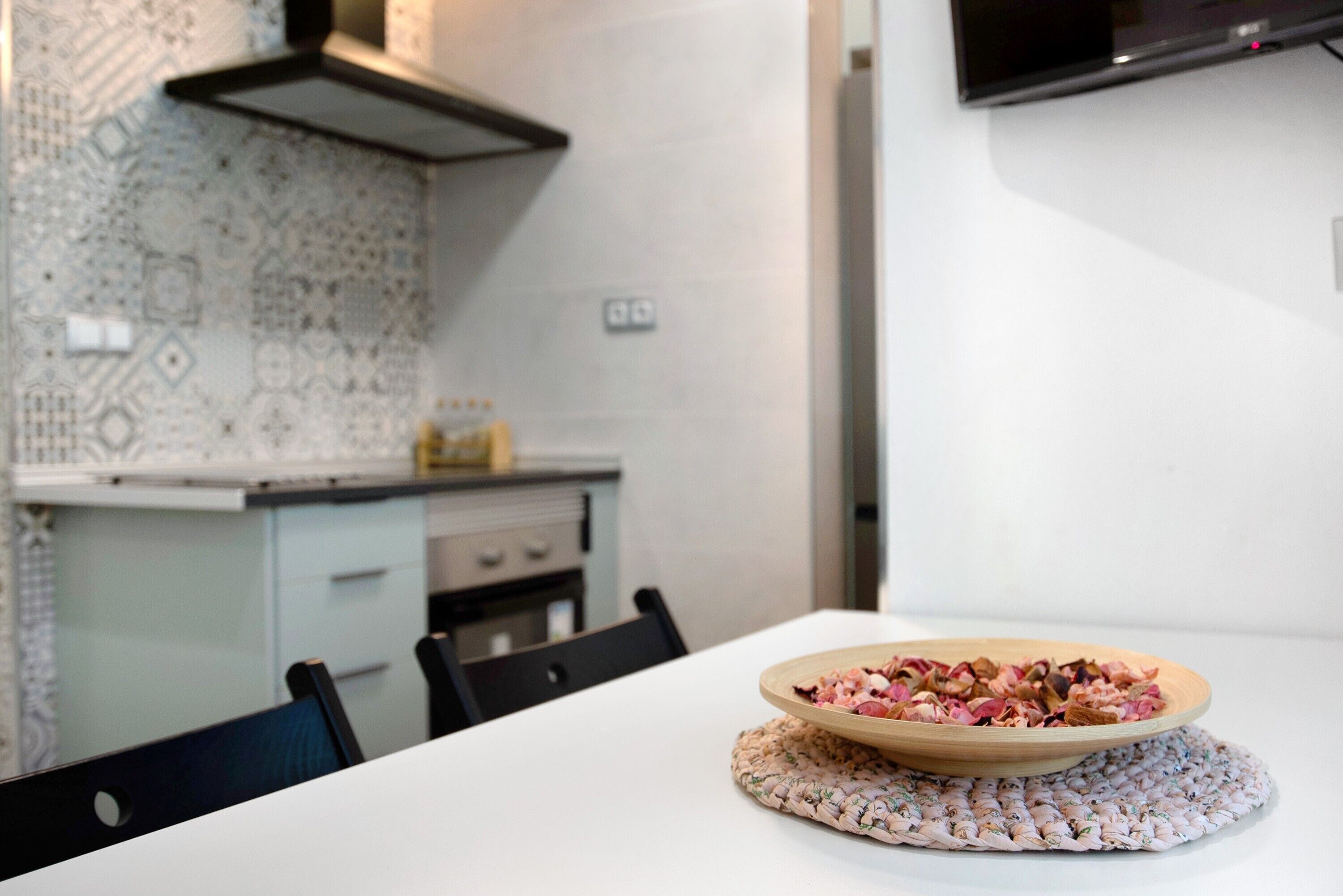 A dining table with a bowl of dried flowers, a woven placemat, and a kitchen in the background.