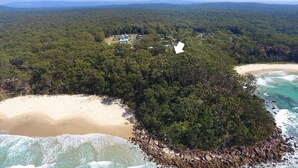 Luchtbeeld van een kustgebied met een strand, dicht bos en een kleine groep gebouwen.