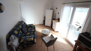 A compact living space with a sofa, a small table, a fan, a TV, a refrigerator, and a sliding glass door leading to an outdoor area. - Charming air-conditioned two-bedroom house with mezzanine, veranda, sleeps five, terrace, WiFi,... (PORTIRAGNES)