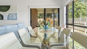 Een moderne eethoek met een glazen tafel en witte, beklede stoelen, een blauwe bank en een groot raam met uitzicht op een houten terras en groen.