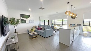 A modern kitchen with a central island, a dining area with chairs, a grey sofa, and a TV on the left.