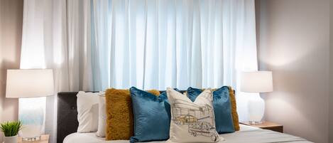 Una cama bien hecha con sábanas blancas, un cabecero y almohadas decorativas. Hay dos mesitas de noche con lámparas y una planta pequeña.