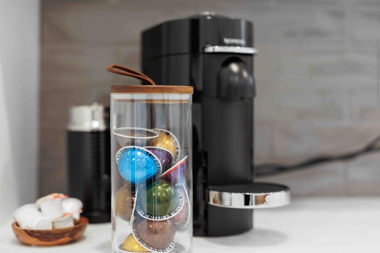A clear glass container with a wooden lid, filled with colorful, round objects, placed in front of a black coffee machine.