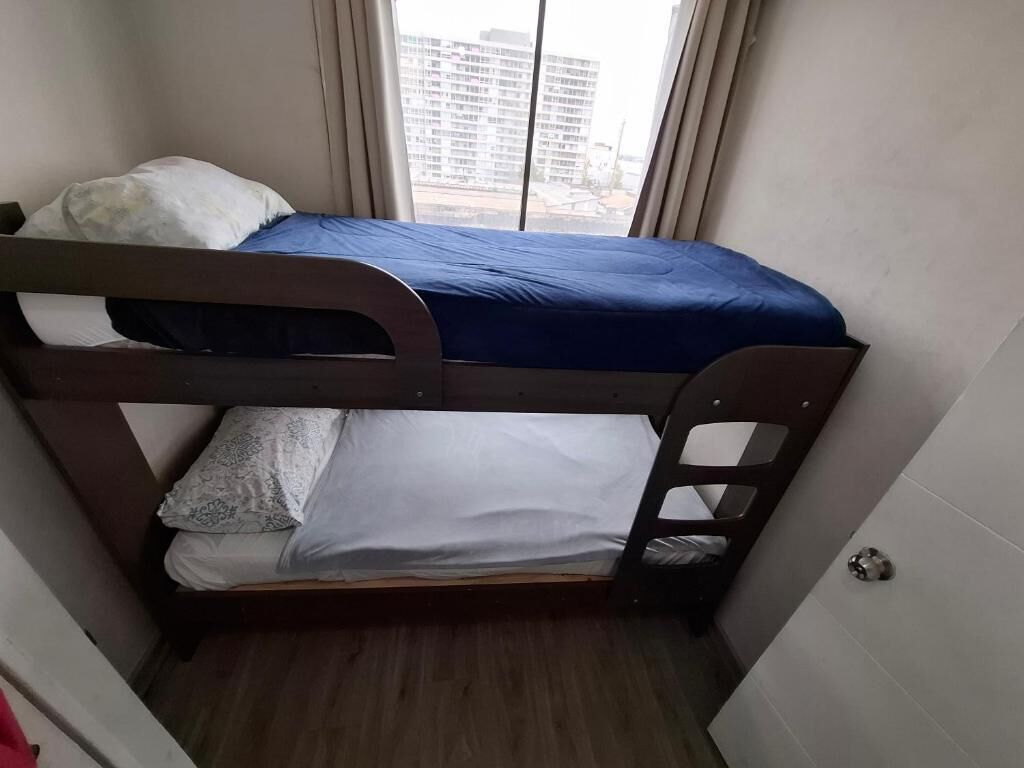 A small, narrow room with a bunk bed, a window with curtains, and a view of a building outside.