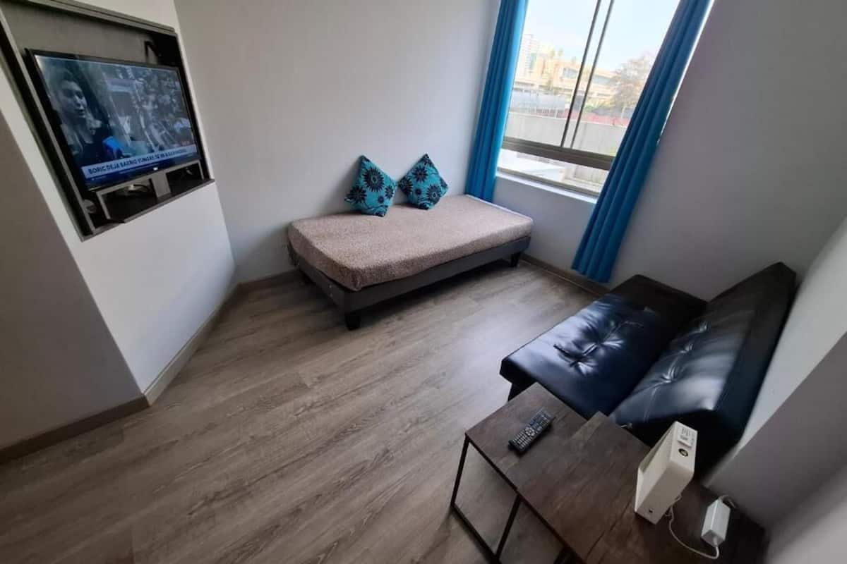 A small living room with a sofa, a small table, a TV mounted on the wall, and a window with curtains.