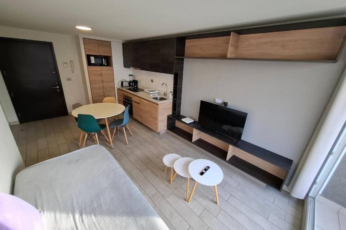 A compact hotel room with a bed, a small dining table with chairs, a kitchenette, a TV, and a sliding door.