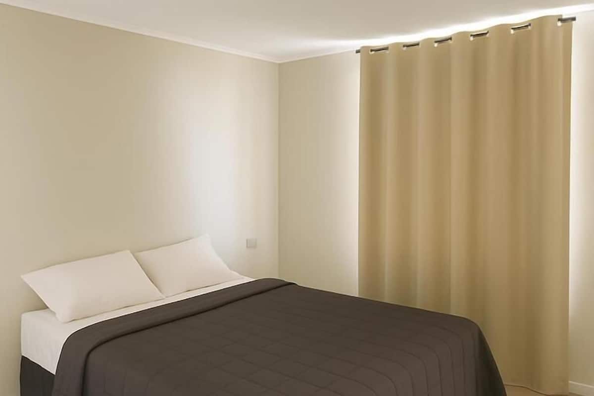 A neatly made bed with a dark-colored bedspread and white pillows, a beige carpet, and a beige wall.