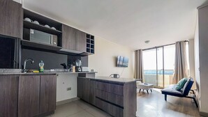 Una cocina moderna con armarios de madera oscura, microondas integrado y una isla grande. Hay un comedor con una mesa y sillas, y un balcón con vistas al mar.