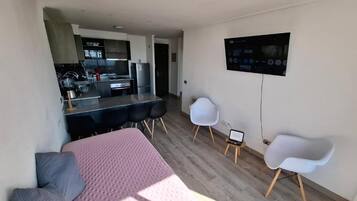 Una habitación moderna con una cama, un escritorio y un televisor. Se ve un área de cocina al fondo.